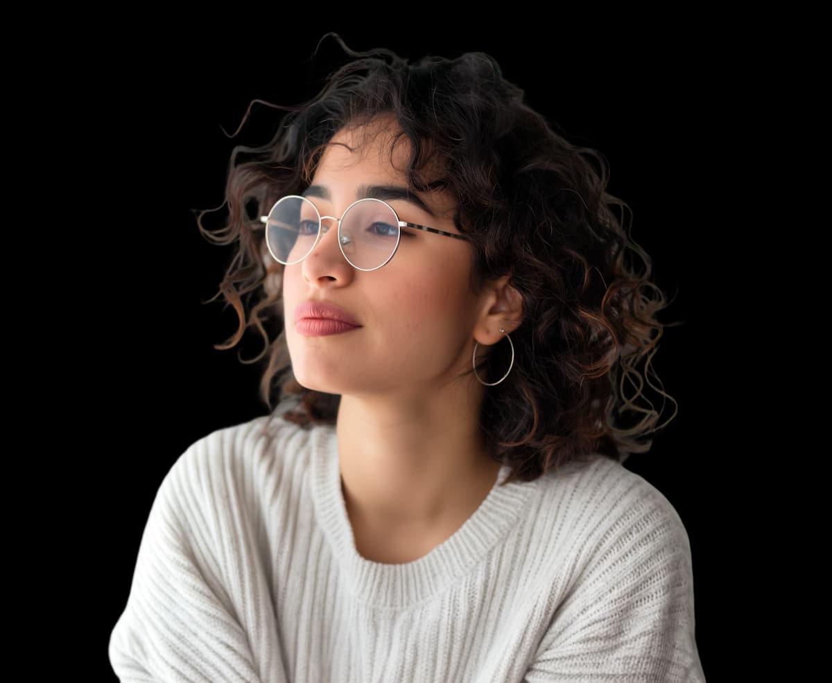 Mulher jovem de cabelos cacheados escuros, usando óculos de armação metálica e suéter branco, olhando pensativa para o lado.